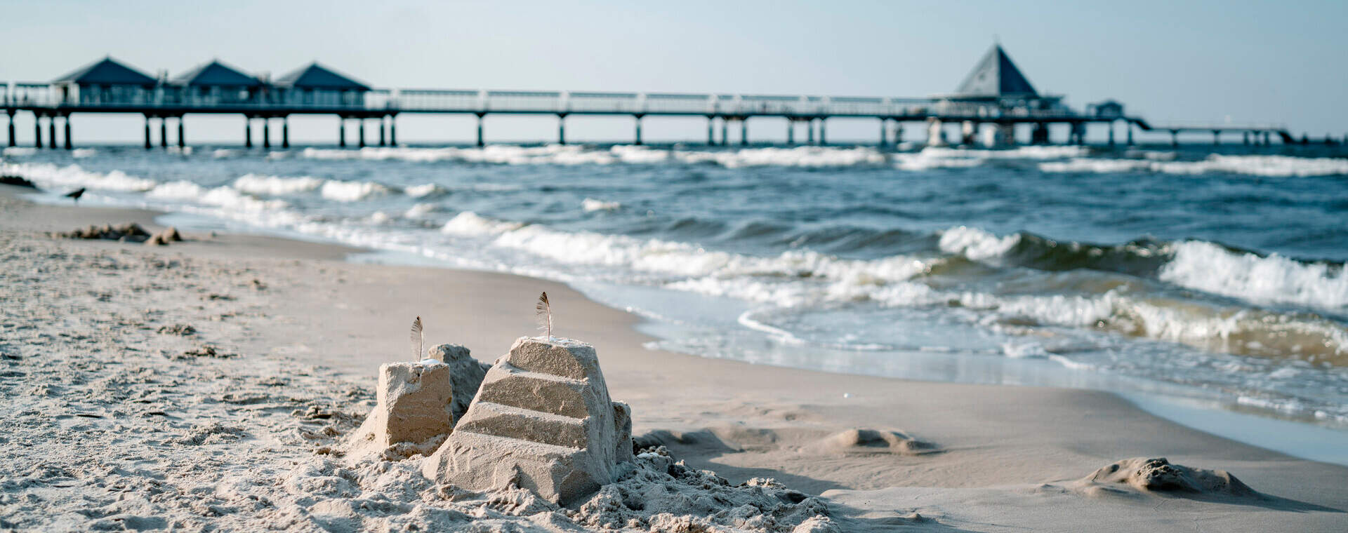 Sandburg am Strand mit Federn, im Hintergrund ein Pier über dem Meer. Sommerlicher Küstenblick.