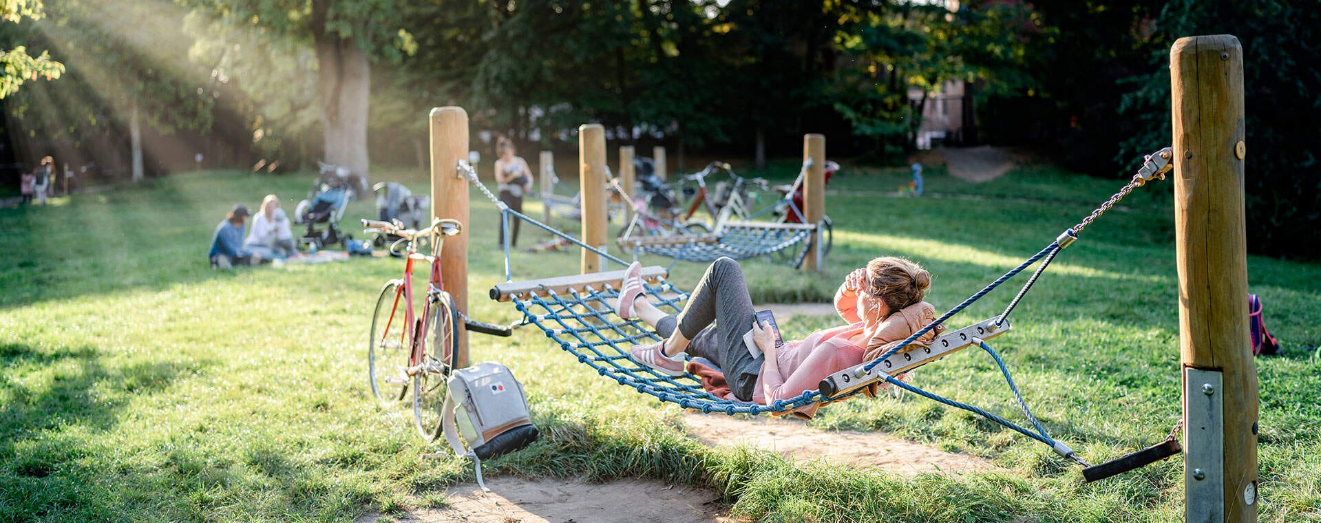 Eine entspannte Person liegt auf einer Hängematte im Park. Fahrräder und Picknickgruppen im Hintergrund.