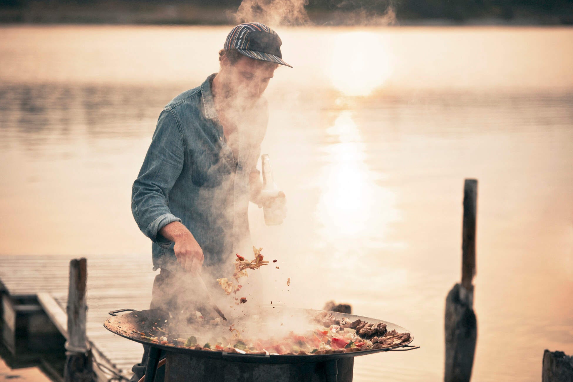 Ein Mann grillt am See, umgeben von Rauch. Entspanntes Kochen in schöner Naturkulisse.